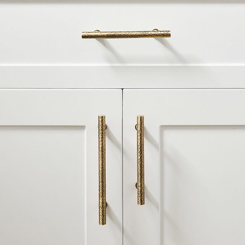 Op de afbeelding: Drie cilindervormige kastgrepen met een getextureerde, gehamerde gouden afwerking bevestigd op witte kastdeuren. E&eacute;n horizontale greep is gecentreerd op de bovenste lade, terwijl twee verticale grepen op de onderste kastdeuren zijn gemonteerd.