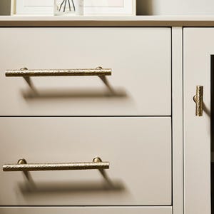 Op de afbeelding: Een lichtbeige kast met twee horizontale lades en &eacute;&eacute;n verticale deur. De lades zijn voorzien van lange, cilindrische handgrepen van gehamerd messing, terwijl de deur een bijpassende verticale, cilindrische knop van gehamerd messing heeft.