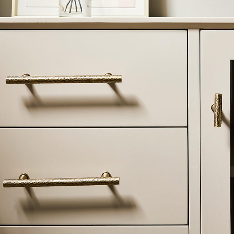 Op de afbeelding: Een lichtbeige kast met twee horizontale lades en &eacute;&eacute;n verticale deur. De lades zijn voorzien van lange, cilindrische handgrepen van gehamerd messing, terwijl de deur een bijpassende verticale, cilindrische knop van gehamerd messing heeft.