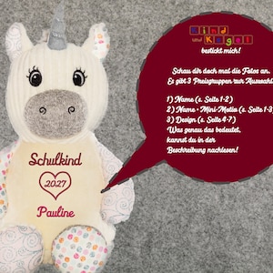 Könnte beinhalten: Ein cremefarbenes Einhorn-Plüschtier mit silbernem Horn und gestickten Details. Das Einhorn hat das Wort "Schulkind" und das Jahr "2027" auf der Brust gestickt, zusammen mit dem Namen "Pauline". Eine Sprechblase enthält deutschen Text.