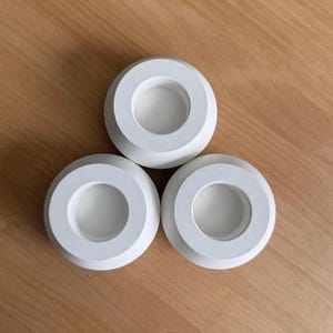 以下が含まれることがあります： 三角形に配置された3つの白い円形のキャンドルホルダー。各ホルダーは滑らかなマット仕上げで、幅の広い平らな縁と、ティーライトキャンドルをしっかりと保持するように設計された中央の深い円筒形の空洞が特徴です。