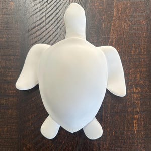 May include: A solid white sea turtle figurine with a smooth, matte finish. The turtle is depicted from a top-down perspective, showing a rounded shell, a head extending forward, two large front flippers, and two smaller rear flippers.
