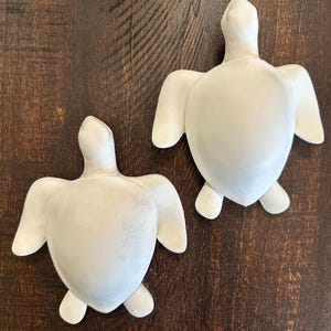 May include: Two matte white sea turtle figurines with smooth, rounded shells and extended flippers. The turtles are positioned with one slightly higher than the other, displaying a uniform, solid color and a clean, minimalist, sculptural form.