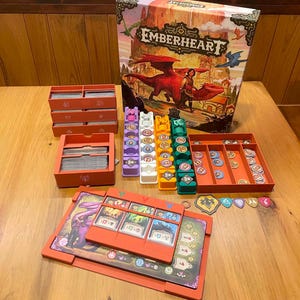 May include: The Emberheart board game set includes a large illustrated box, orange card storage trays, and four vertical token organisers in purple, white, orange, and teal. It features a player board with orange card slots and various circular game tokens.