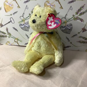 May include: A yellow plush teddy bear with a pink ribbon around its neck. The bear has a Ty Beanie Babies tag attached to its ear. The bear is sitting on a white fabric surface.