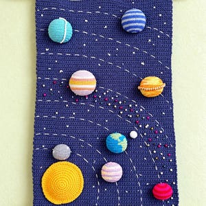 Op de afbeelding: Een donkerblauwe gehaakte wandhanger met een zonnestelselontwerp, voorzien van diverse kleurrijke, bolvormige, gevulde planeten die op het oppervlak zijn bevestigd. Witte gestikte lijnen vormen banen, geaccentueerd door kleine, verspreide, veelkleurige kralen.