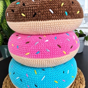 Peut inclure: Une pile de trois coussins ronds en forme de beignet crochetés. Le coussin du haut est marron avec des vermicelles colorés, celui du milieu est rose vif avec des vermicelles, et celui du bas est bleu vif avec des vermicelles.