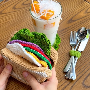 May include: A crocheted sandwich toy featuring tan bread slices, a white egg, a red tomato slice, a yellow cheese slice, and a ruffled green lettuce leaf. Beside it sits a glass of mango smoothie and a set of metal cutlery tied with green yarn.