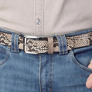 May include: A belt with a snakeskin pattern in shades of beige, gray, and black, featuring a silver-toned metal buckle. The belt is threaded through the loops of blue denim jeans, worn with a light gray button-down shirt tucked into the waistband.