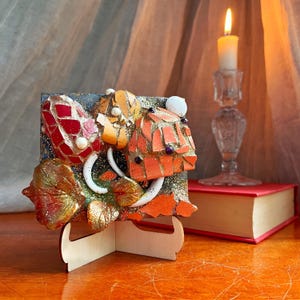 May include: A square mosaic art piece featuring red, orange, and gold glass shards with white accents on a glittery dark background. The artwork is displayed on a light-colored wooden stand, showcasing textured leaf shapes and small beads.