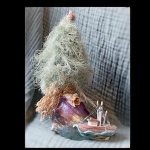 May include: A whimsical, handmade nautical-themed art piece. It features a small, detailed boat with masts and rigging, set against a backdrop of a purple and gold structure, and a fluffy, green tree-like element. The piece is set on a blue fabric.