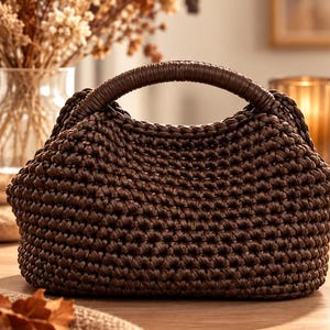 Puede incluir: Un bolso de mano marrón oscuro elaborado con una textura de cordón grueso tejido a ganchillo. El bolso presenta un asa integrada redondeada, envuelta en un material liso marrón oscuro a juego, creando una silueta semicircular y estructurada.