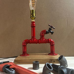 May include: A table lamp constructed from red painted metal pipes mounted on a rectangular wooden base. The lamp features a brass socket with an Edison-style light bulb and a small black metal faucet handle attached to the pipe structure.