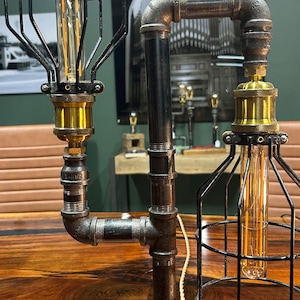 May include: A dual-light table lamp constructed from dark grey industrial metal pipes mounted on a rectangular wooden base. The lamp features two vertical Edison-style bulbs, each enclosed within a black wire cage and accented with brass sockets.