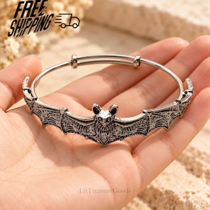 May include: A silver-toned metal bangle bracelet featuring a central bat motif with outstretched wings. The bat design has intricate, textured detailing on the wings and body, and the bracelet has a thin, adjustable circular band structure.