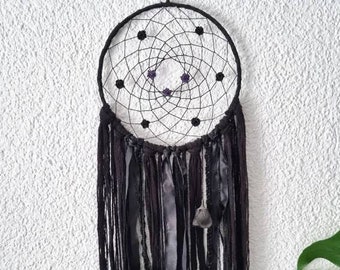 May include: A black dreamcatcher with purple beads and black feathers hanging from the bottom. The dreamcatcher is hanging from a white wall.