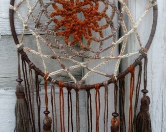 May include: A handcrafted dreamcatcher with a circular frame wrapped in brown yarn. The web features a central orange sunburst design and is woven with beige, brown, and tan yarn. Brown tassels and beads hang from the bottom, creating a bohemian aesthetic.