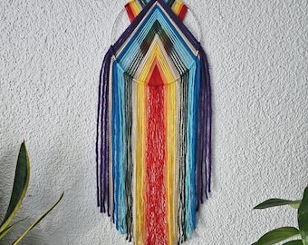 Puede incluir: Un colorido tapiz de pared hecho a mano con un diseño de arcoíris. La pieza presenta una forma de V central con hebras verticales de hilo en rojo, amarillo, azul y morado. Un aro plateado y una cuerda para colgar en la parte superior. Un artículo decorativo.
