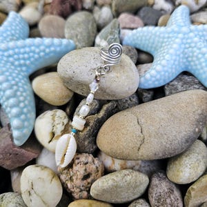 May include: A silver-toned ear cuff with a spiral design, adorned with a dangling strand of beads, pearls, and a white seashell. The cuff rests on a stone, with light blue starfish and other stones in the background. A unique piece of jewelry.