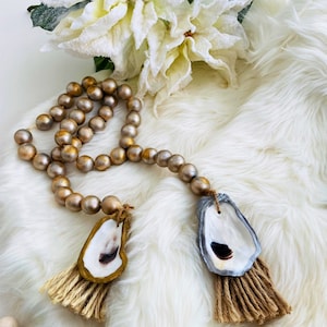 May include: A gold and silver beaded garland with two oyster shell ornaments with brown tassels. The garland is on a white faux fur background.