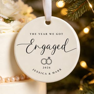 May include: A round white ceramic ornament hanging from a white ribbon. The text reads THE YEAR WE GOT Engaged, followed by an illustration of two interlocking rings and the year 2026, with the names JESSICA & MARK printed at the bottom.