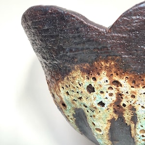 May include: A ceramic bowl with a dark brown, textured rim and a porous, cratered surface featuring a blend of beige, light green, and black glaze. The bowl has an irregular, wavy edge and a small, narrow base, showcasing a rustic finish.
