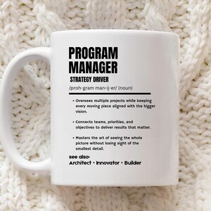 May include: A white ceramic mug featuring black printed text that defines a Program Manager as a strategy driver. The text includes a phonetic pronunciation, a noun classification, three bulleted job descriptions, and a see also section.