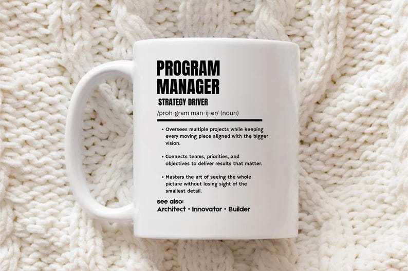 May include: A white ceramic mug featuring black printed text that defines a Program Manager as a strategy driver. The text includes a phonetic pronunciation, a noun classification, three bulleted job descriptions, and a see also section.