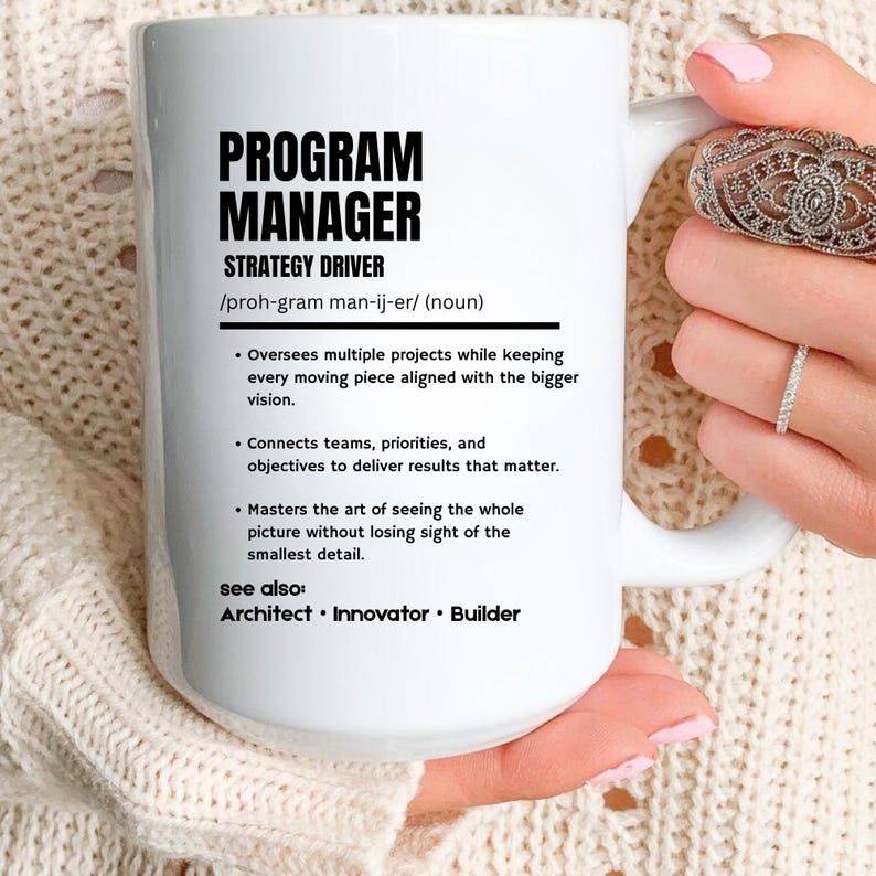 May include: A white ceramic mug featuring black printed text that defines a Program Manager as a strategy driver. The text includes a phonetic pronunciation, three bulleted job responsibilities, and a see also section listing Architect, Innovator, and Builder.