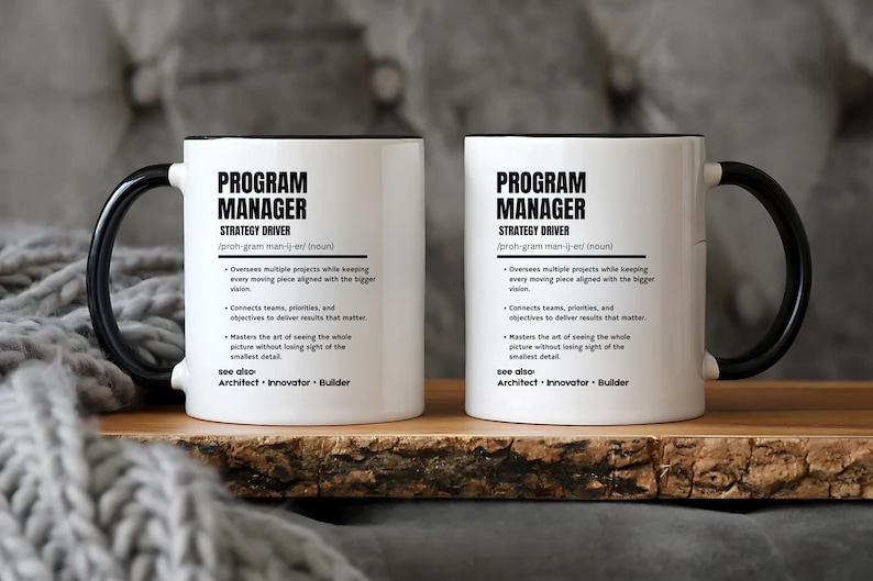 May include: Two white ceramic mugs with black handles and rims, each featuring black printed text that defines a program manager as a strategy driver, including bulleted job responsibilities and a see also section listing architect, innovator, and builder.