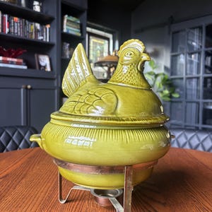 May include: A glazed olive green ceramic chicken-shaped tureen with a lid, featuring a textured feather pattern on the body. The tureen rests on a metallic stand with three legs and a central circular base, designed for holding a heat source.