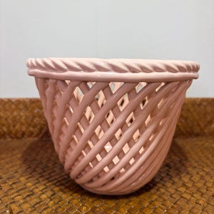 Peut inclure: Un panier en céramique rose clair avec une finition brillante, présentant un motif en treillis tressé sur le corps et une bordure épaisse en forme de corde torsadée le long du rebord supérieur. La structure est ouverte et arrondie.