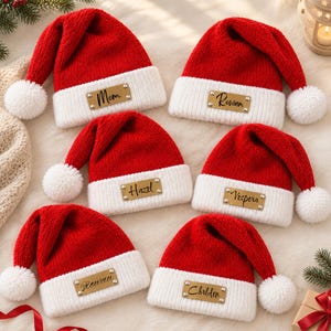 May include: Six red and white knit Santa hats, each featuring a white pom-pom at the tip and a rectangular gold-toned metal tag attached to the white cuff. The tags are engraved with the names Mom, Rowan, Hazel, Vespera, Rowan, and Chrlden.