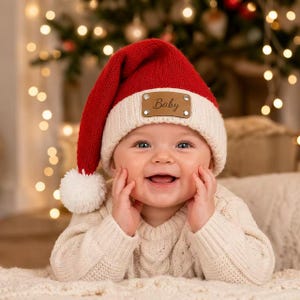 May include: A red and white knit hat with a pom-pom and a rectangular brown leather patch featuring the word Baby in cursive script. The hat has a ribbed white cuff and a soft red crown, designed to be worn on the head during the holiday season.