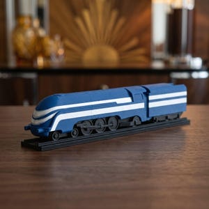 Könnte beinhalten: Eine blau-weiße Modelleisenbahn-Lokomotive mit stromlinienförmigem Design, horizontalen weißen Streifen am Gehäuse, schwarzen Rädern und einem schwarzen Sockel, präsentiert als statisches Sammlerstück auf einer ebenen Fläche.
