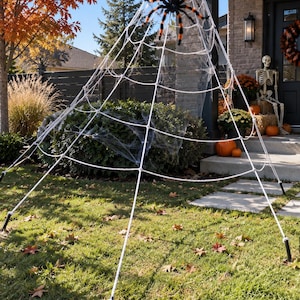 May include: A large white rope spider web decoration anchored to the ground with black stakes, extending upwards to a roof edge. A large black spider with orange accents is attached to the centre of the web, creating a tall, conical display.