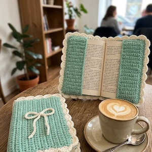 May include: A mint green crocheted book cover with a cream-coloured scalloped border and a decorative cream bow on the front. A second matching cover is open, holding an open book, positioned next to a ceramic mug filled with latte art coffee.