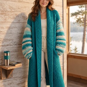 Könnte beinhalten: Eine lange, vorne offene Häkelstrickjacke in einfarbigem Petrol mit langen, weiten Ärmeln, die horizontale Streifen in Petrol, Hellblau und Creme aufweisen. Das Kleidungsstück hat einen breiten Schalkragen und zwei tiefe Vordertaschen.