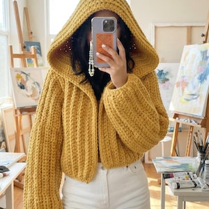 Puede incluir: Un cárdigan con capucha de color amarillo mostaza, tejido en punto grueso con textura acanalada y mangas largas. La prenda presenta una capucha grande y puntiaguda con un diseño abierto en la parte frontal. El tejido es grueso y prominente.