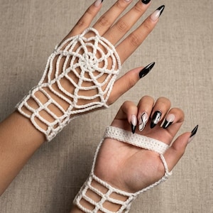 Puede incluir: Un par de guantes sin dedos de ganchillo blanco con un patrón de telaraña en el dorso de la mano y un diseño de rejilla simple en la muñeca. Los guantes están confeccionados con hilo blanco y dejan los dedos y el pulgar al descubierto.