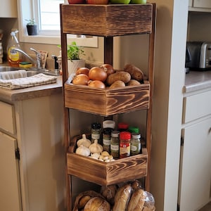 May include: A four-tier corner storage rack made of dark stained wood with a scorched finish. The shelves hold various items including red and green apples, yellow onions, brown potatoes, garlic bulbs, assorted spice jars, and loaves of bread.