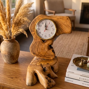May include: A table clock featuring a round white face with black Arabic numerals and a gold-tone bezel, set into a natural, irregular piece of light brown wood with visible grain patterns and knots, standing on a matching wooden base.