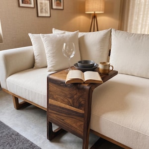 May include: A C-shaped side table crafted from dark brown wood with a visible grain pattern, positioned over a cream-colored sofa. The table surface holds an open book, a clear glass wine glass, a dark gray bowl, and a two-toned ceramic mug.