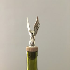 Peut inclure: Un bouchon de bouteille argenté orné d'un aigle aux ailes déployées. Le bouchon est monté sur un bouchon de liège et est placé sur une bouteille en verre vert. L'aigle a des plumes détaillées.