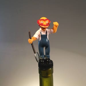 May include: A yellow cartoon figure with red hair and a blue jumpsuit holding a shovel, standing on top of a wine bottle stopper.