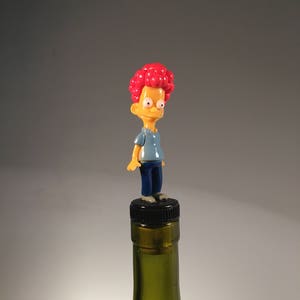 May include: A novelty bottle stopper featuring a cartoon character with red hair, a blue shirt, and blue trousers. The figure is standing on a black bottle cap, atop a green glass bottle. The stopper adds a touch of fun to any wine bottle.
