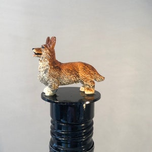 May include: A brown and white dog figurine with a black nose and tongue, standing on a black bottle stopper. The bottle stopper is made of wood and has a black finish.