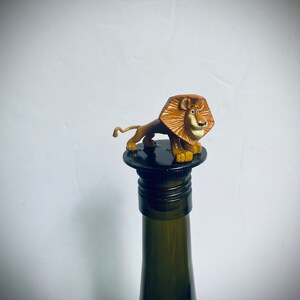 May include: A brown lion figurine with a black nose and blue eyes is on top of a black wine bottle stopper. The bottle is green glass.