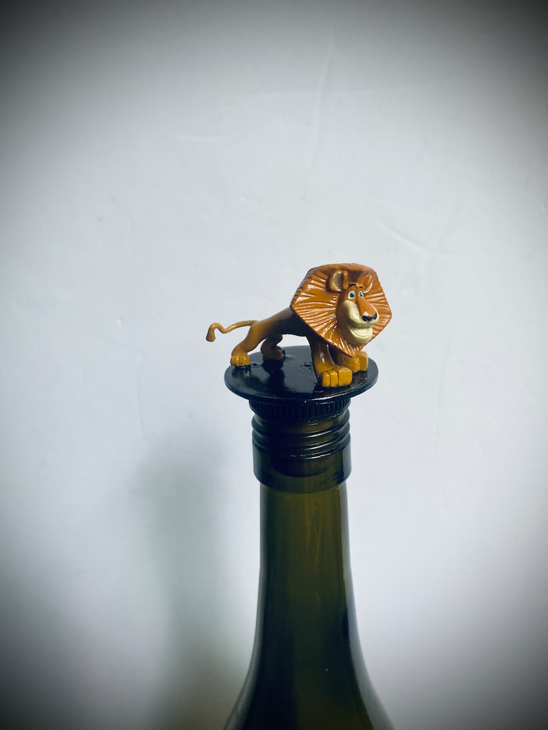 May include: A brown lion figurine with a black nose and blue eyes is on top of a black wine bottle stopper. The bottle is green glass.