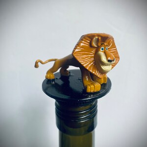 May include: A brown lion figurine with a blue eye and a black nose is sitting on top of a black bottle stopper. The stopper is on top of a green glass bottle.